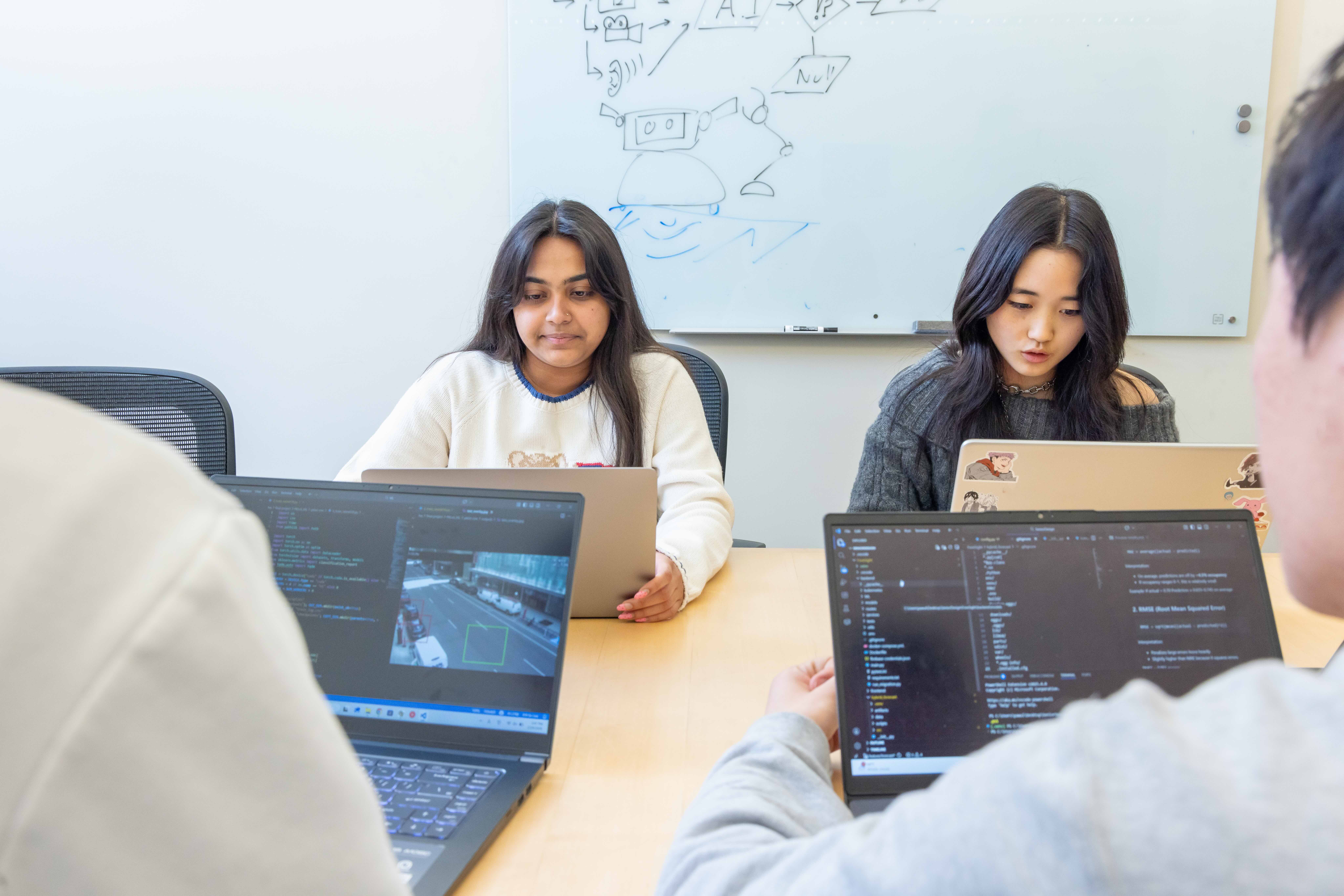 Two women code at their computers, framed between two computers running code